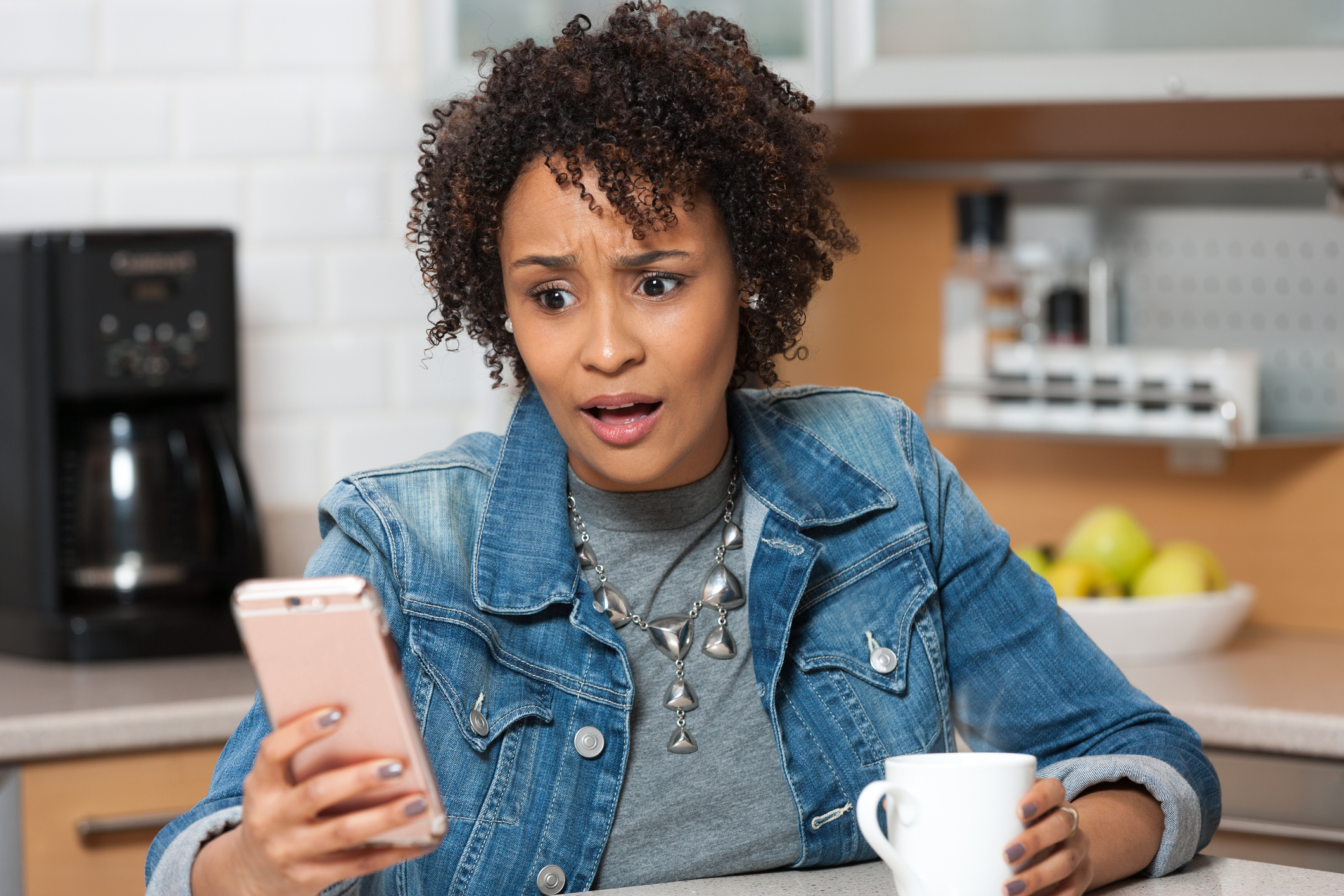 Young African American woman on mobile phone with shocked look on her face, sitting in kitchen