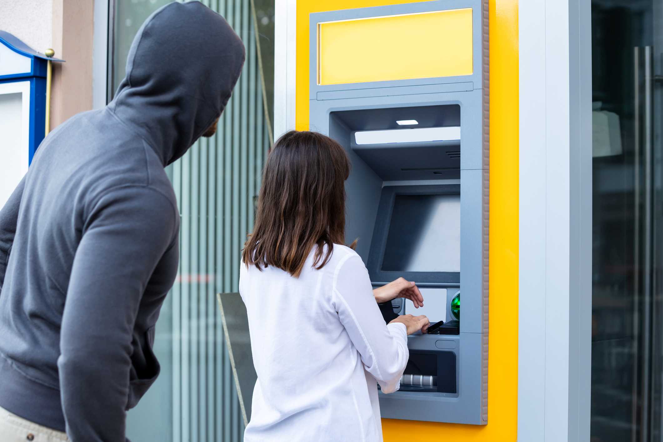 Woman using an outdoor bank ATM as a man in a hoodie behind her tries to see her PIN