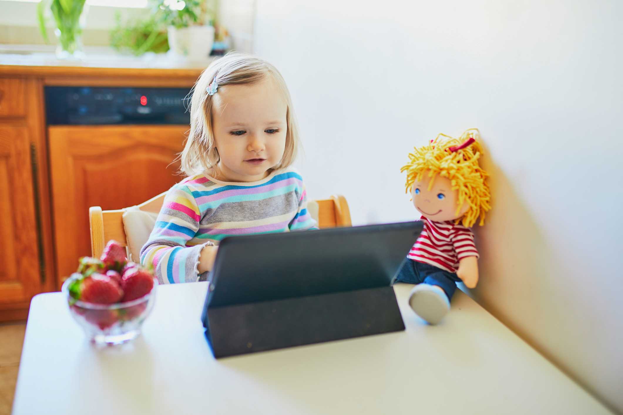 A small little girl looking at a tablet computer screen as she sits at a small table in a home kitchen. A bowl of strawberries and a rag doll are also on the table.