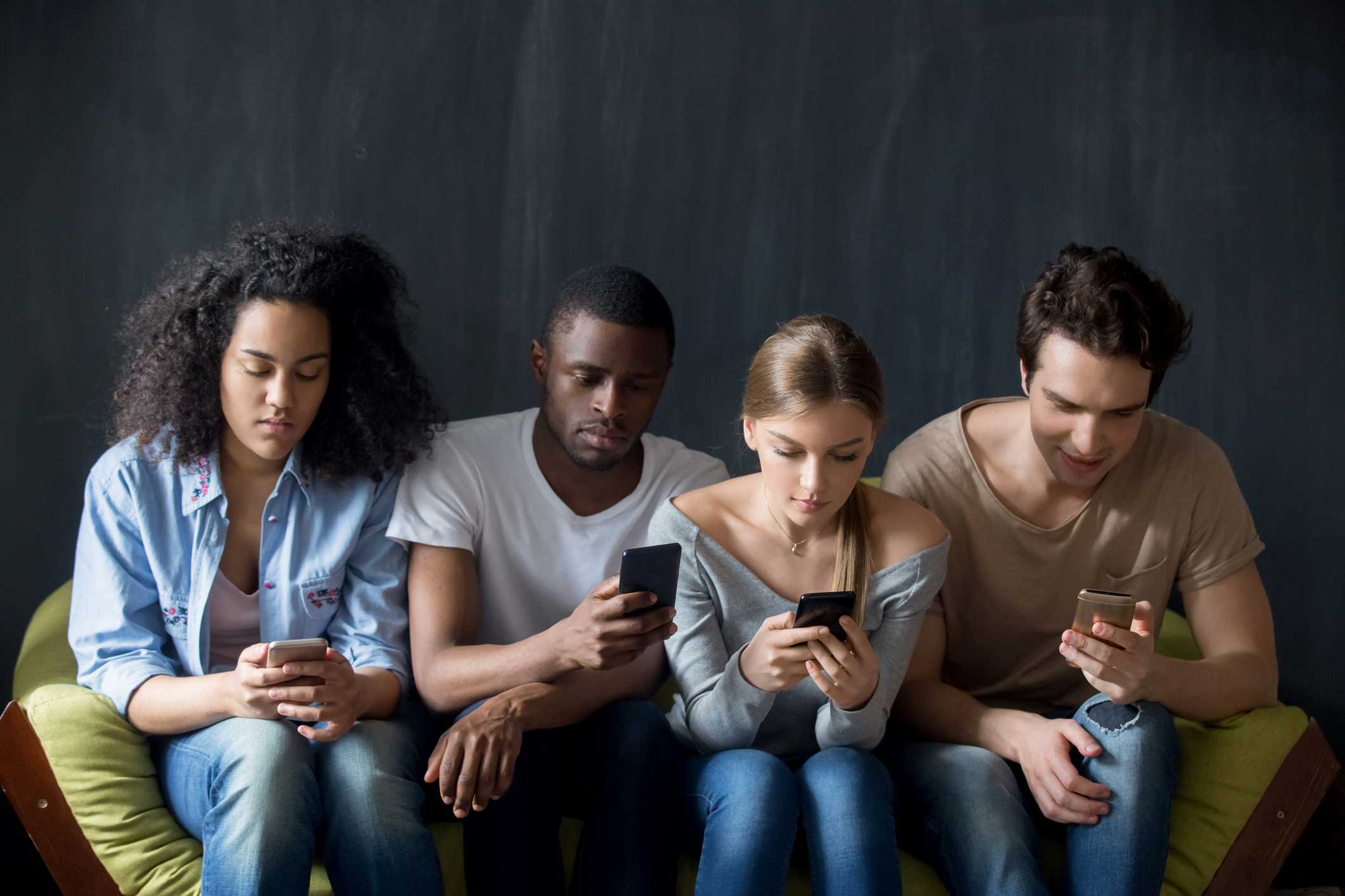 4 young people sitting on a sofa and looking at their mobile phone screens.