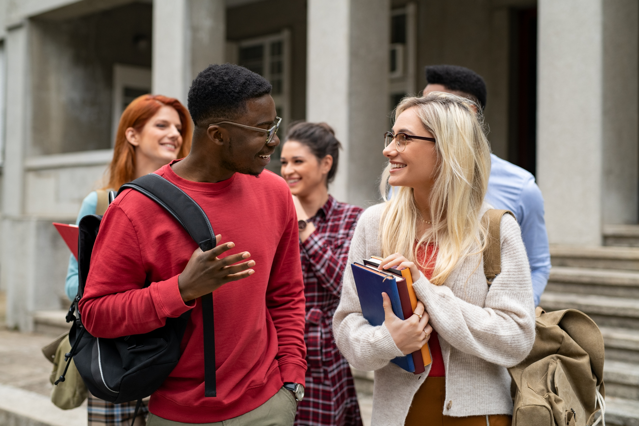 Group of happy multiethnic students walking outside a college building. Five university students holding books and school backpacks while talking together.