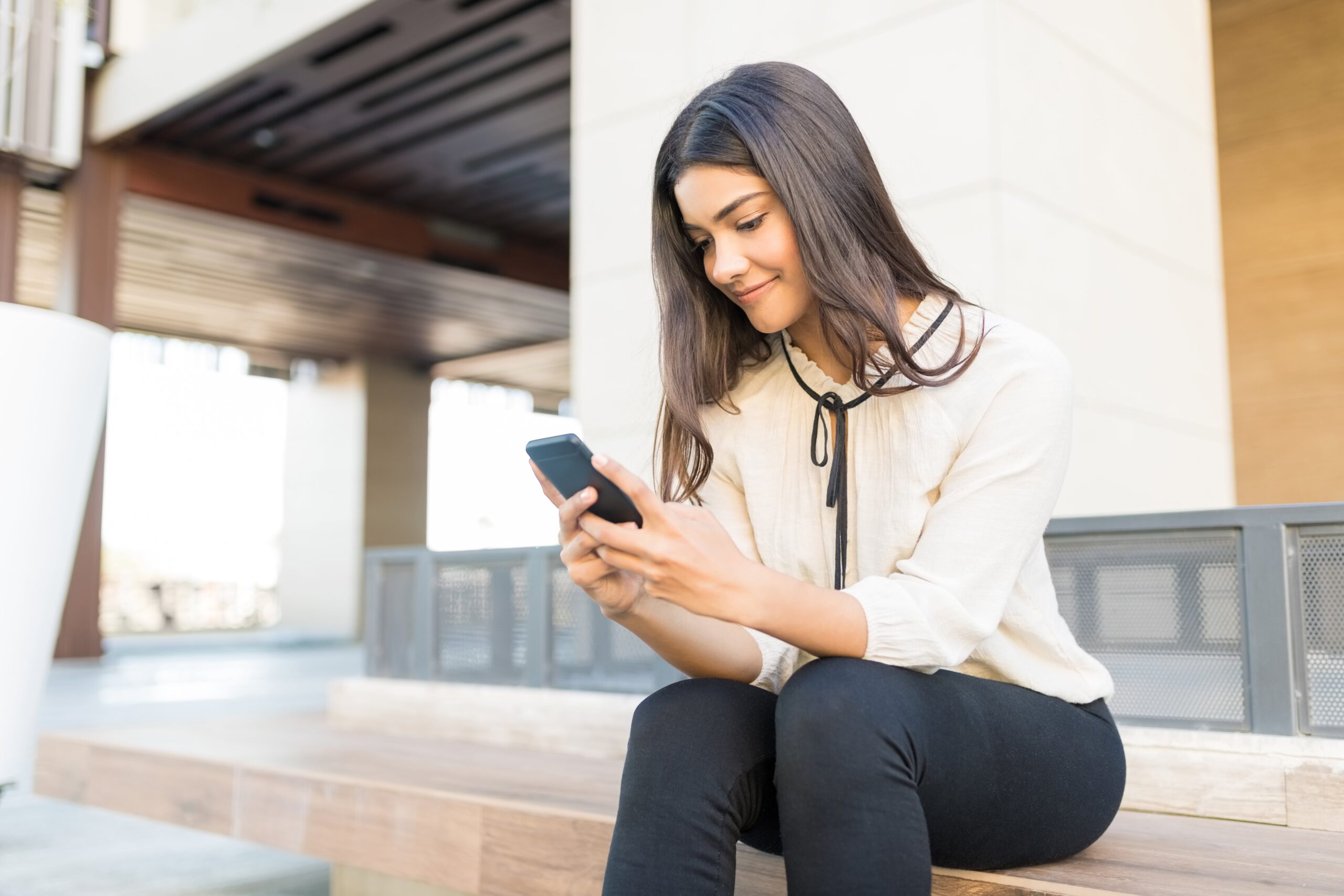 Young woman sitting on office building bench looking at her mobile phone.