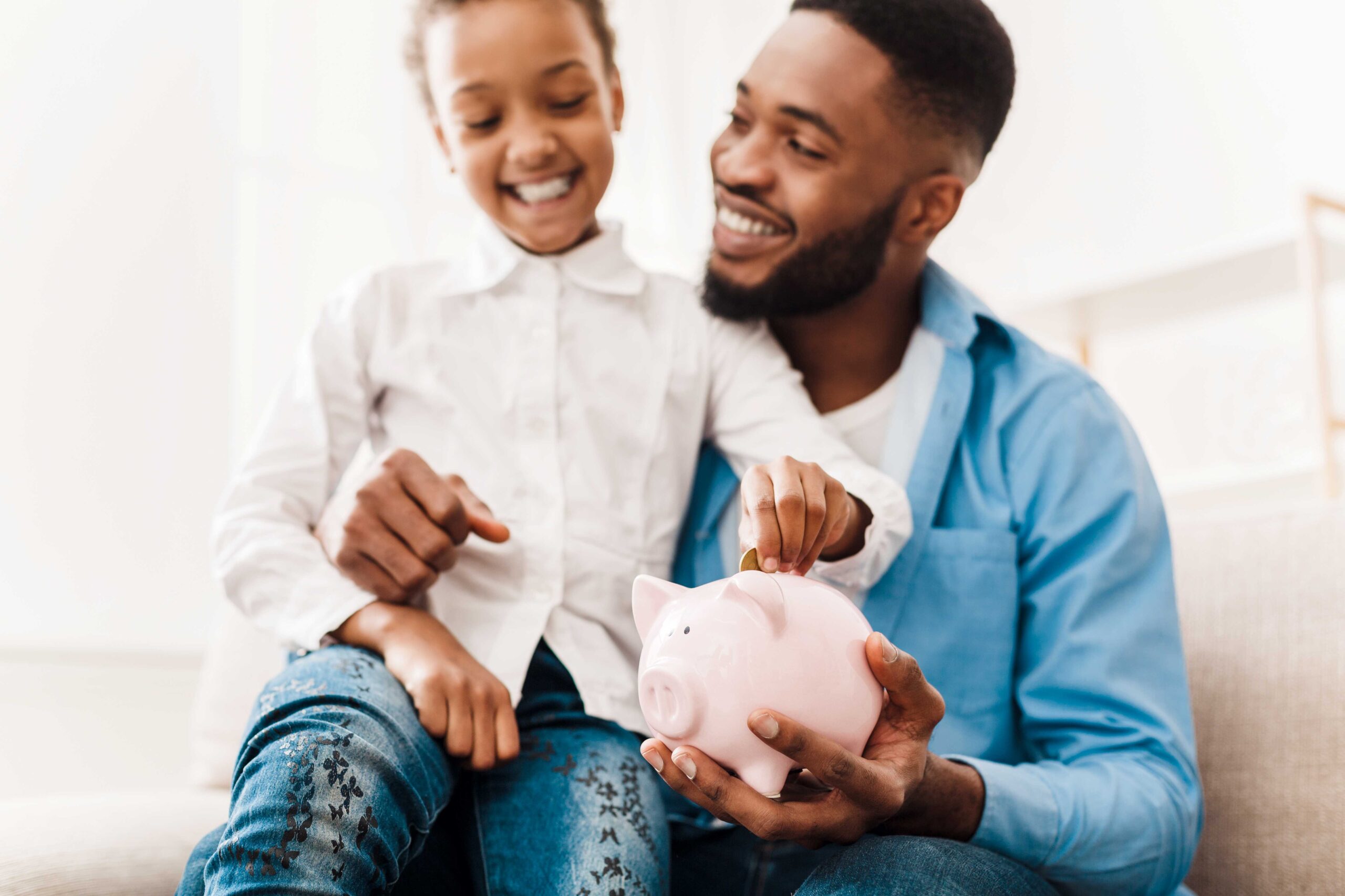 A man holding a young girl who is placing coins into a pink piggy bank.