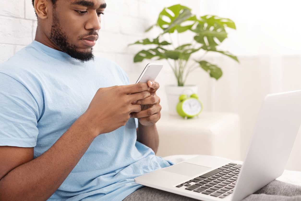 A man relaxing on a bed, typing on a smartphone, and holding a laptop.