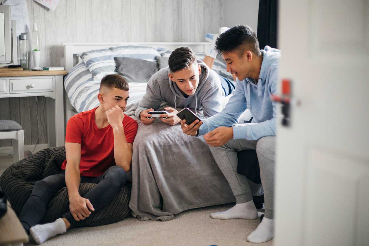 Three teenage boys looking at social media on a mobile phone screen while relaxing in a bedroom.