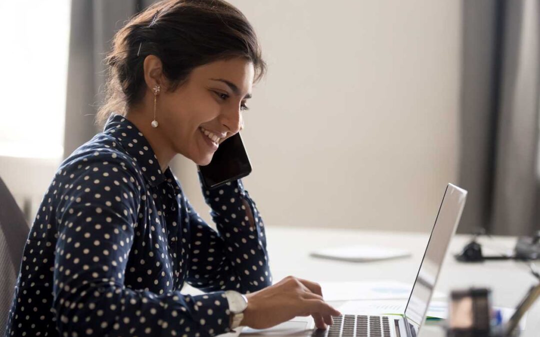 Smiling business woman talking on a mobile phone while typing on a laptop.