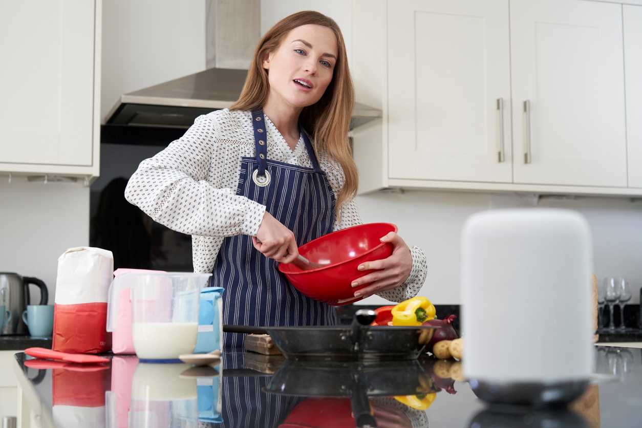 Woman in a kitchen stirring ingredients in a red bowl as she looks at a white smart speaker.