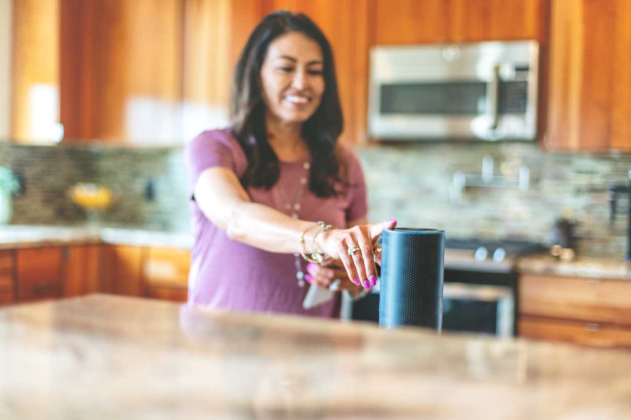 A woman standing in a home kitchen touches a smart speaker.