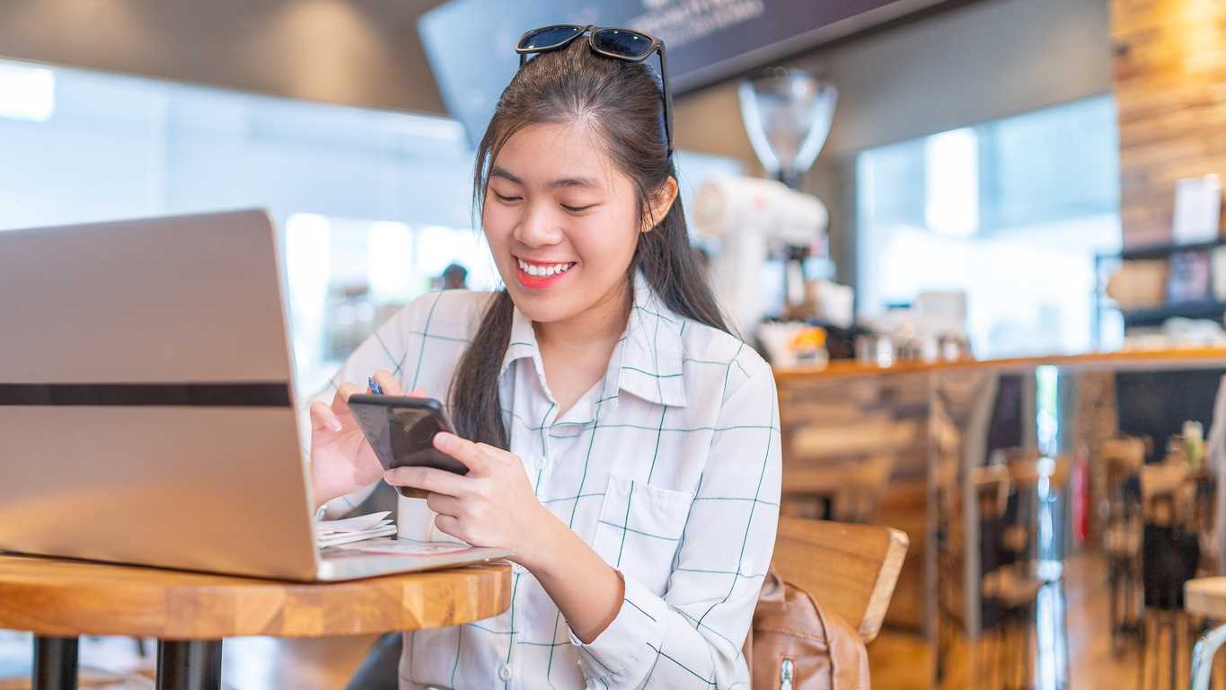 A woman working in a coffee shop. She smiles as she navigates her smartphone.