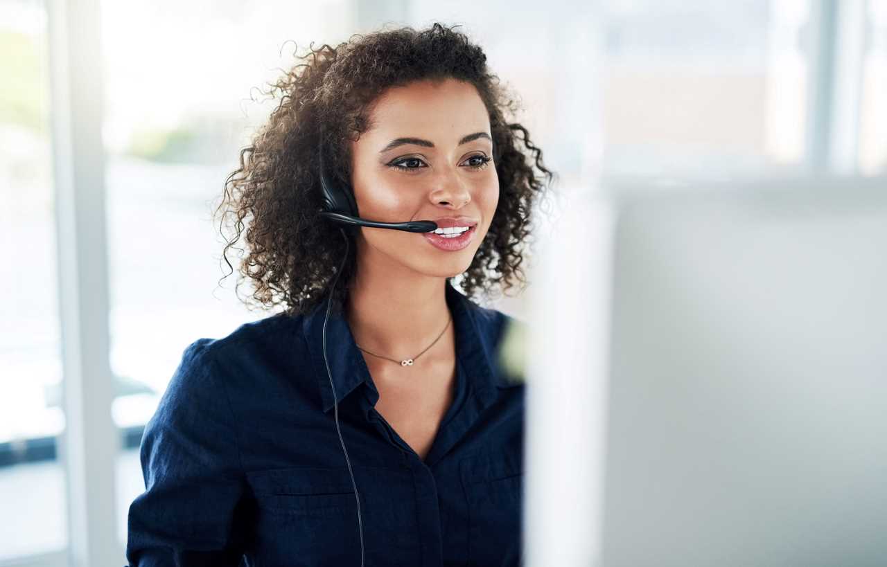 A female customer service agent wearing a headset and looking at a computer monitor as she talks to a customer.