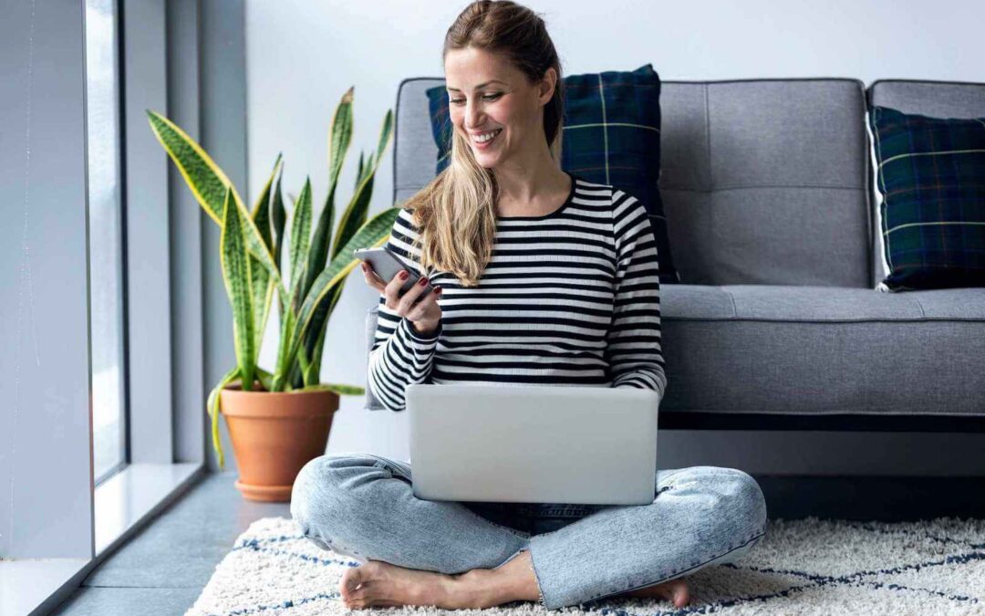 Smiling woman sitting on the floor in front of a sofa with a laptop and mobile phone.