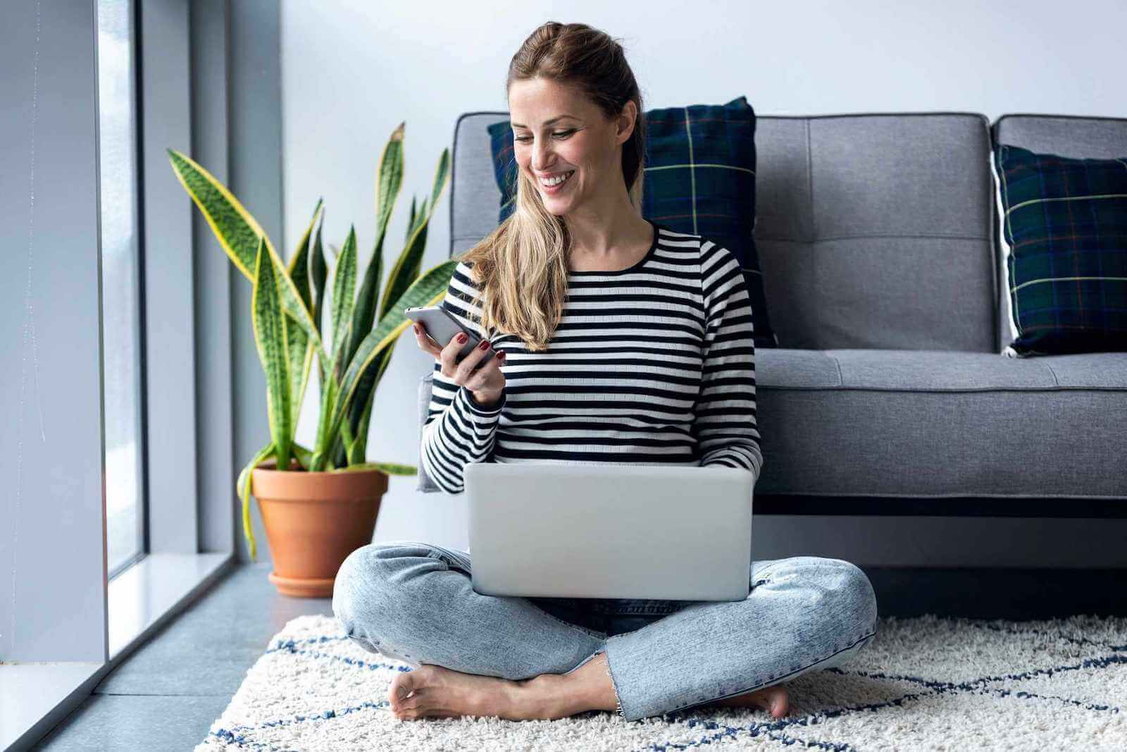 Smiling woman sitting on the floor in front of a sofa with a laptop and mobile phone.