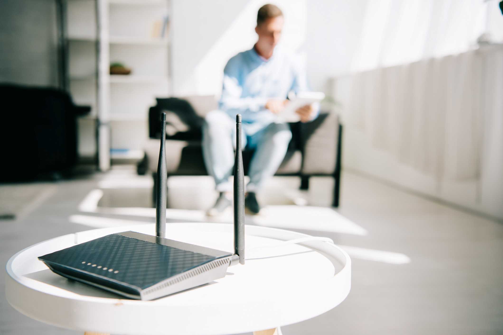 Home Wi-Fi modem on a small, white, round table. A man is using a tablet computer in the background.