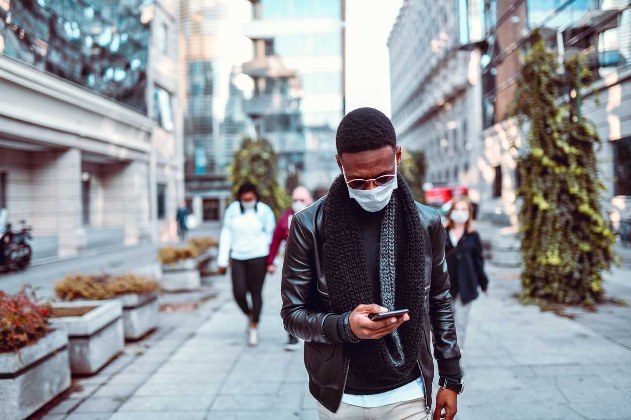 A man wearing a face mask walks down a city sidewalk as he looks at his smartphone.