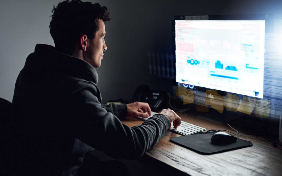 A hacker sitting at a desk and viewing data on a large computer screen.