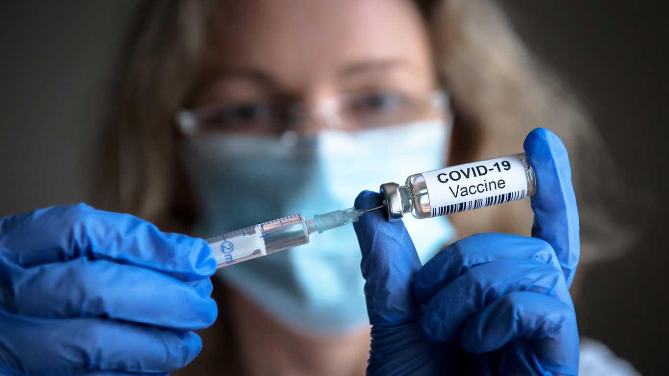 A medical worker fills a syringes with the COVID-19 Vaccine. She's wearing a blue face mask and blue latex gloves.