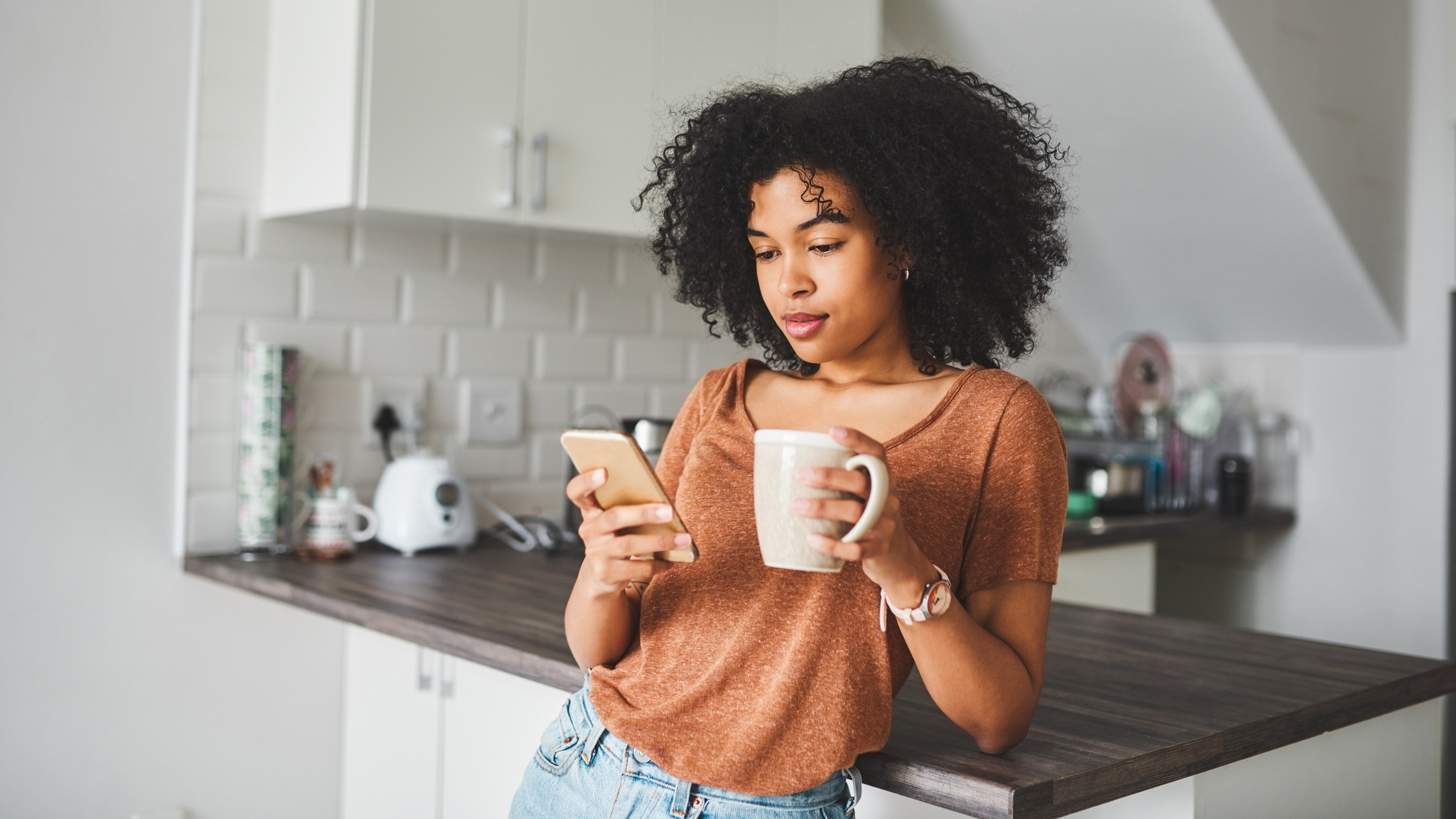 Young woman holding and viewing a smartphone while having coffee in the kitchen at home