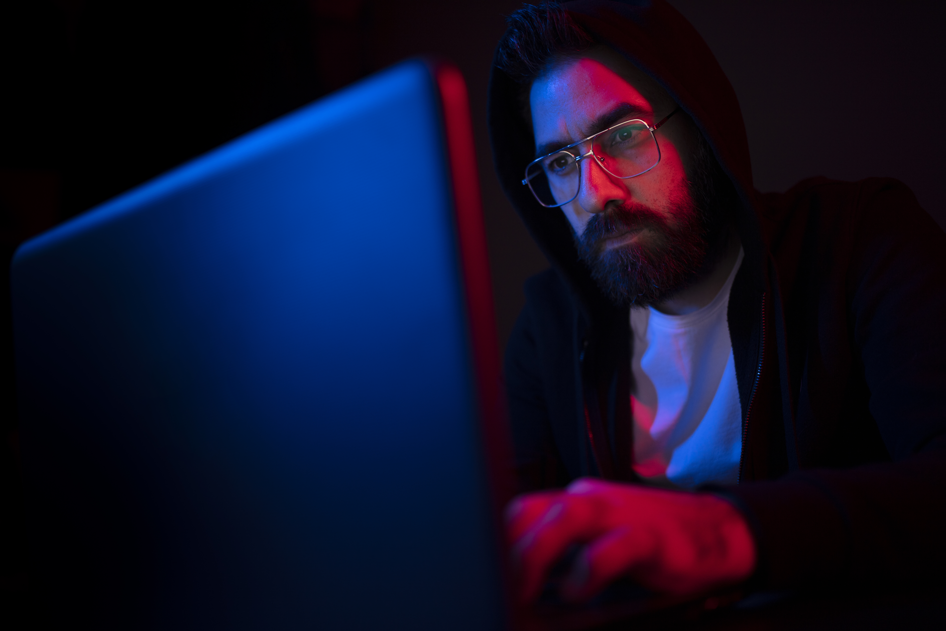 Man with facial beard and wearing eyeglasses and a black hoodie types on a laptop in a dark room.