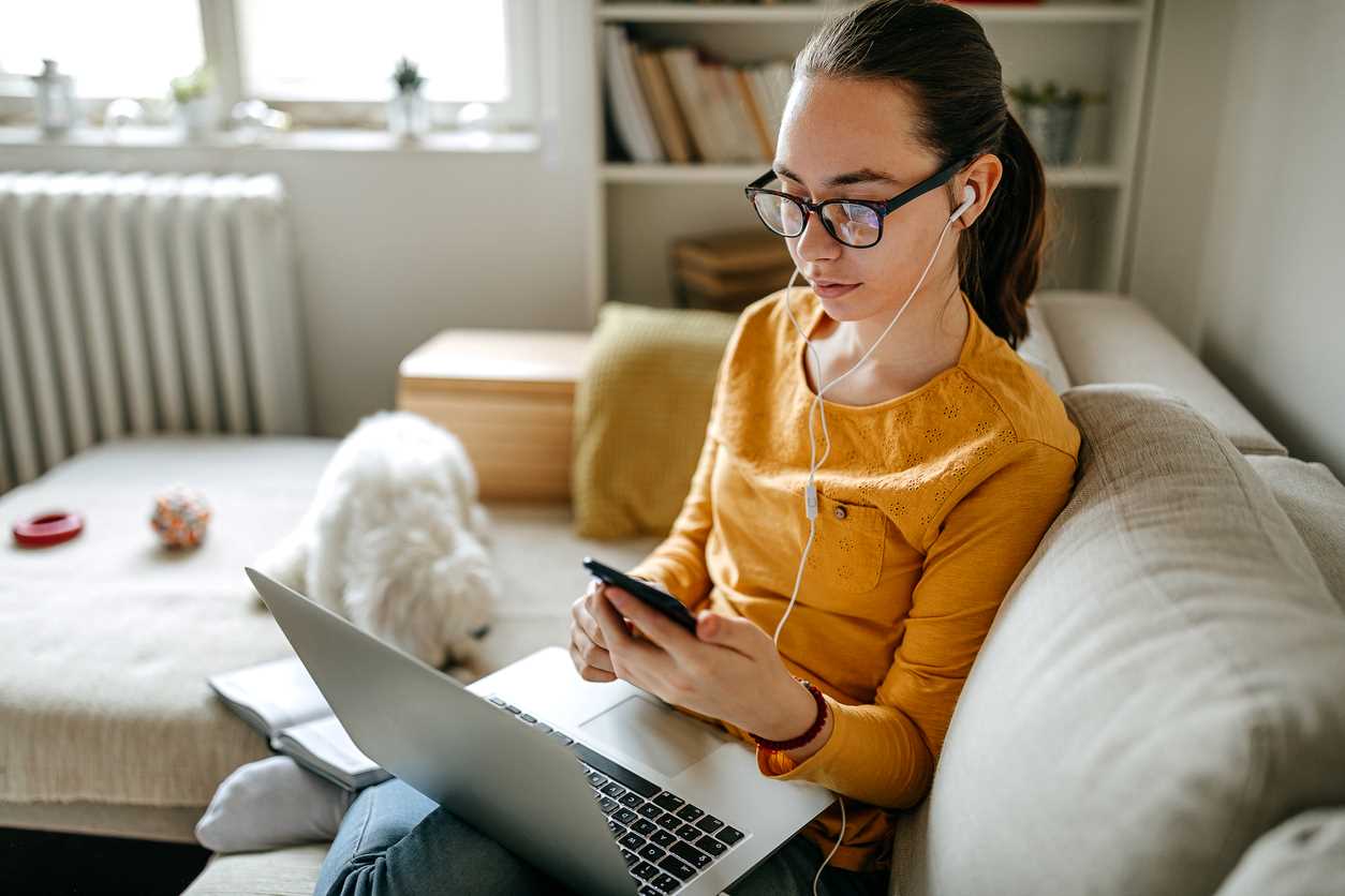 Woman casually sitting on her sofa, looking at mobile phone and holding a laptop. She is wearing earphones and a small white dog plays nearby on the sofa.