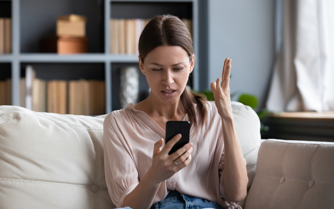 A frustrated woman sitting on a living room sofa looking at her smartphone.