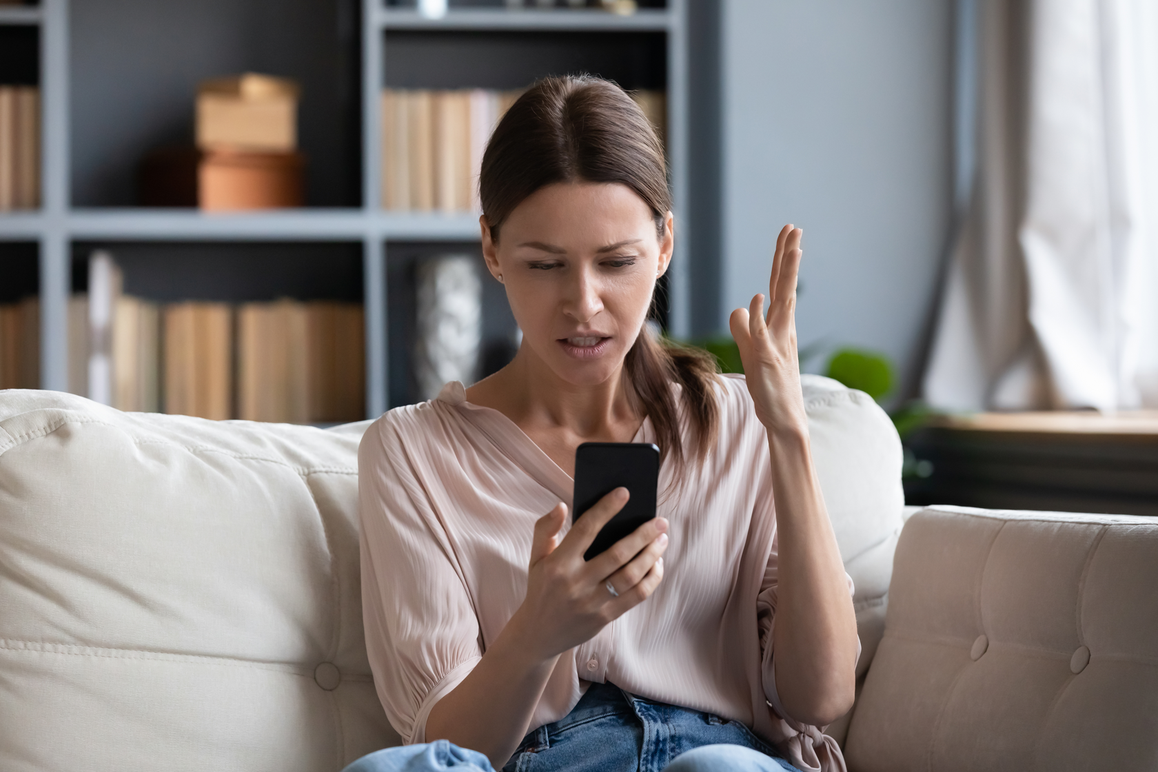 A frustrated woman sitting on a living room sofa looking at her smartphone.