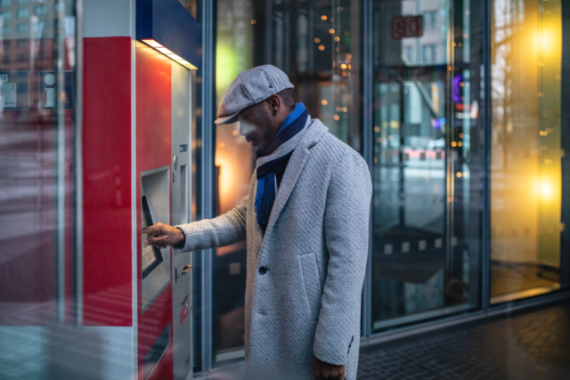 Man using an outside ATM. He's dressed in a winter coat and hat.