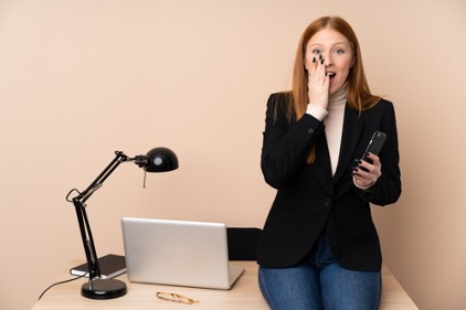Young professional women leaning against her work desk with her hand to her face realizing she needs to delete info from her social media accounts.