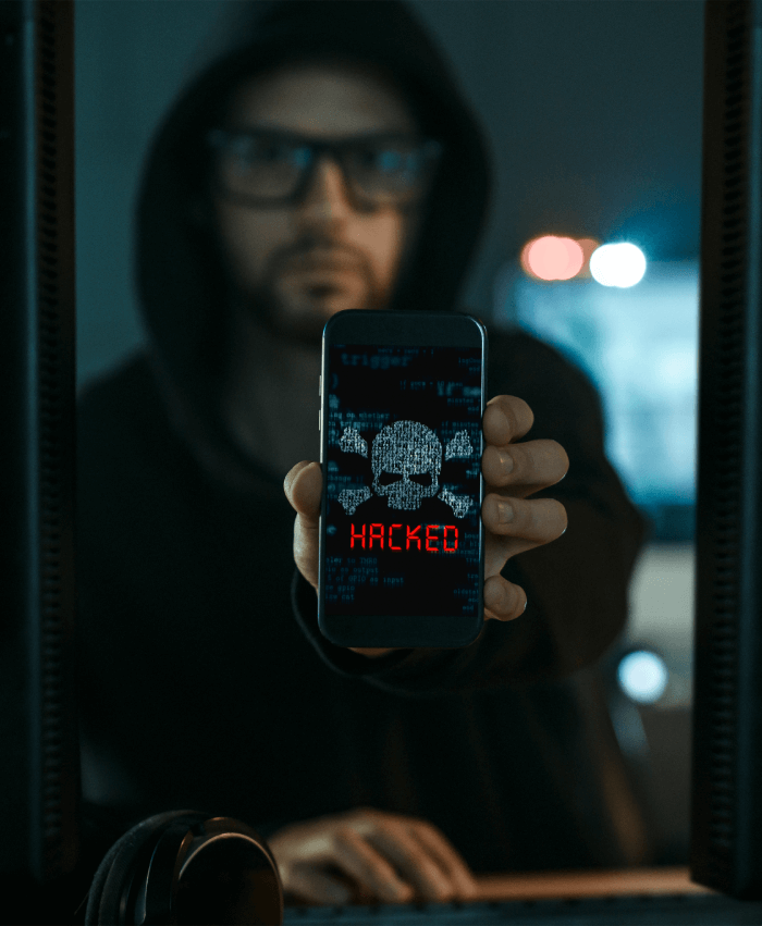 Man in a black hoody holding up a phone with cross bones and skull above the word Hacked.