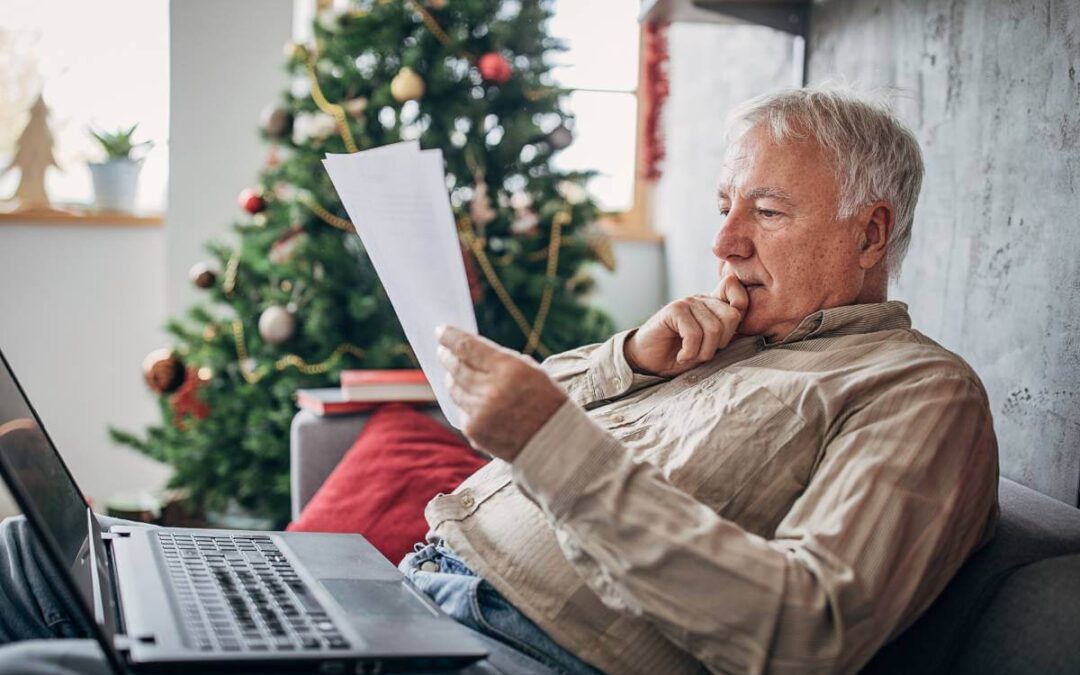 An older man sitting on a sofa looking at his credit report. A laptop and Christmas tree is nearby.