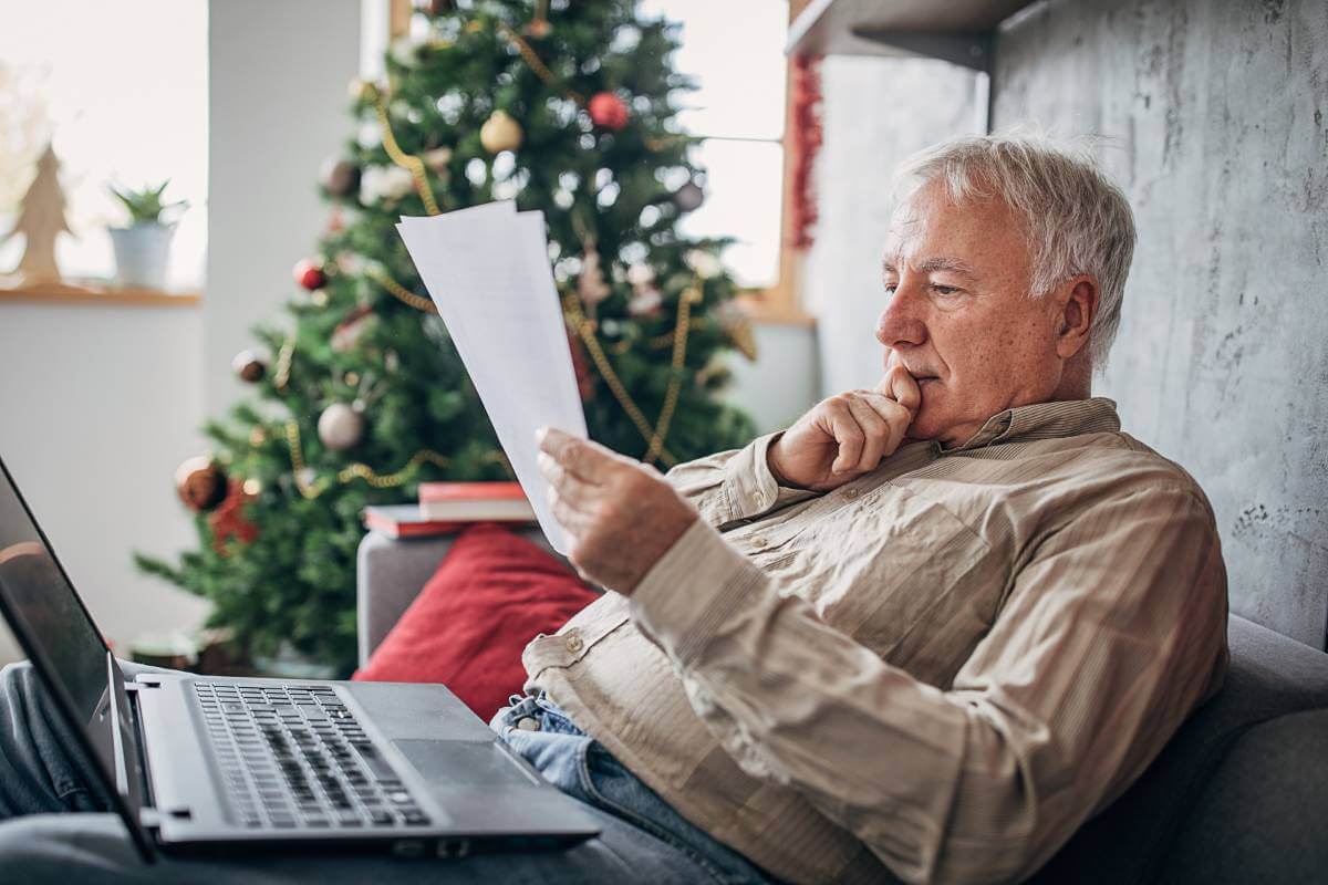 An older man sitting on a sofa looking at his credit report. A laptop and Christmas tree is nearby.