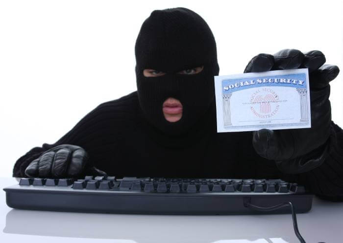 Thief in a face mask holding a Social Security Card as he types on a computer keyboard.